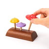 A hand interacts with a Wooden Mushroom Spinning Top Set featuring three colorful mushrooms in red, yellow, and purple, set on a dark brown base made of responsibly-sourced wood. The red mushroom is being picked up.