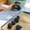 A hand forms small round seed balls from soil, placing them on parchment paper as part of the DIY Seed Ball Kit - Rainbow Garden STEM activity. A green container and a brick wall can be seen in the background.