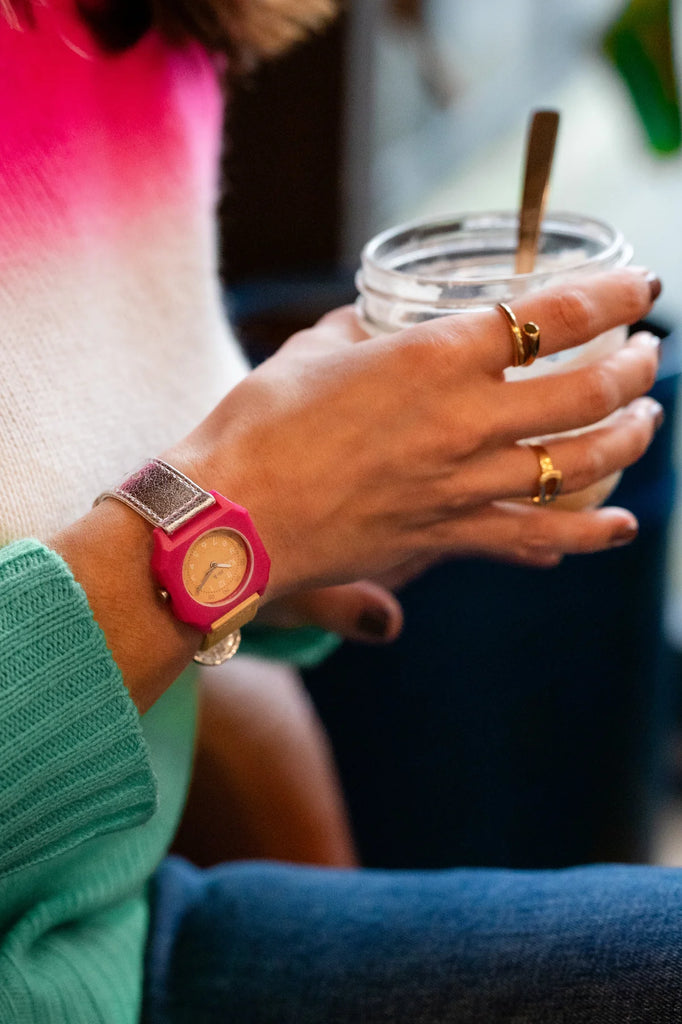 A person wearing a pink Mini Kyomo Watch - Disco and gold rings holds a glass jar with a metal spoon, dressed in a green sweater and a white and pink gradient top.