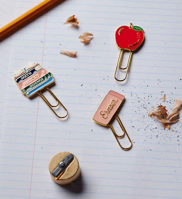 The Schoolhouse Enamel Paper Clip Set, featuring three decorative clips shaped like a red apple, stacked books, and an eraser, rests on lined notebook paper beside pencil shavings, a pencil, and a sharpener.