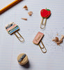 The Schoolhouse Enamel Paper Clip Set, featuring three decorative clips shaped like a red apple, stacked books, and an eraser, rests on lined notebook paper beside pencil shavings, a pencil, and a sharpener.