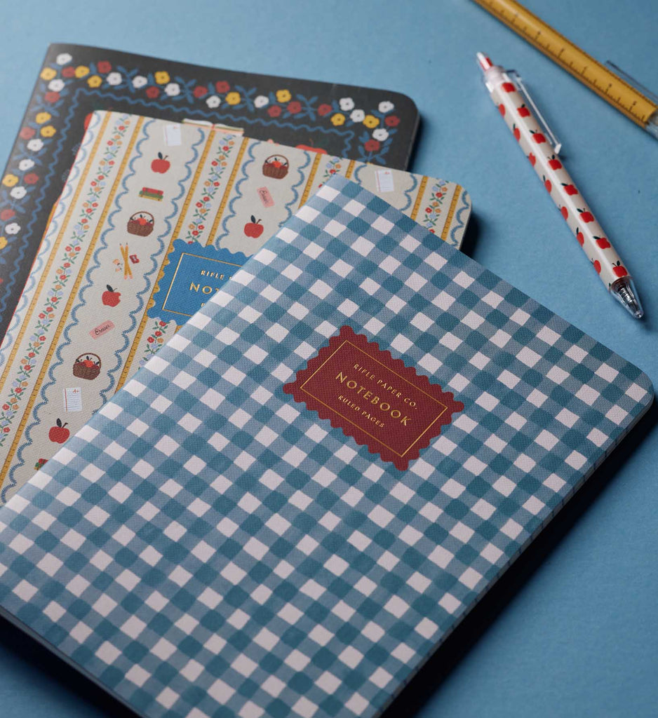 Three patterned notebooks from the Assorted Set of 3 Schoolhouse Notebooks, a white pen decorated with red hearts, and a yellow ruler are arranged on a blue surface. The top notebook showcases a blue and white checkered cover with a red label.