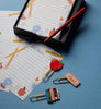 The Schoolhouse Note Writing Set features lined paper adorned with school-themed illustrations, a red pen, and three novelty paper clips shaped like an apple, an eraser, and stacked books—all arranged on a blue surface—making it perfect for any note-writing collection.