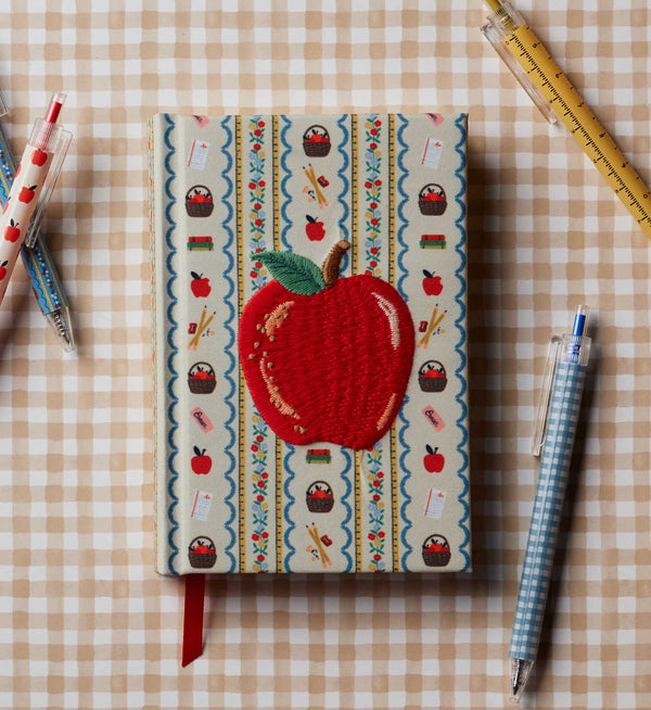 The Schoolhouse Mini Embroidered Journal, a pocket-sized hardbound notebook with a fabric cover, is embroidered with a red apple and decorated with delightful apple and cupcake patterns, pictured amidst pens and a pencil on a beige checkered background.