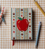 The Schoolhouse Mini Embroidered Journal, a pocket-sized hardbound notebook with a fabric cover, is embroidered with a red apple and decorated with delightful apple and cupcake patterns, pictured amidst pens and a pencil on a beige checkered background.