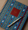 The 2027 Schoolhouse 17-Month Academic Hardcover Spiral Planner, featuring school-themed illustrations and a gold spiral binding, rests on a beige surface. This large blue planner is paired with a decorative pen capped in blue, placed diagonally across its cover, offering both weekly and monthly planning in style.