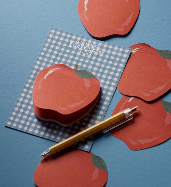 Apple-shaped die-cut sticky notes decorate a blue gingham notepad, with more adhesive notepads scattered around and a yellow pen resting on the blue surface.