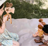 A young girl sits on a blanket outdoors, using the Father’s Factory Super 16 Pro Wooden Toy Camera with Tripod to film two dolls. The pretend-play scene unfolds on grass, bathed in natural light.