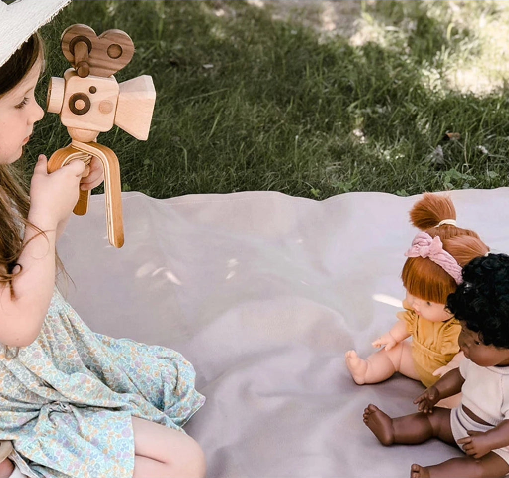 A young girl sits on a blanket outdoors, using the Father’s Factory Super 16 Pro Wooden Toy Camera with Tripod to film two dolls. The pretend-play scene unfolds on grass, bathed in natural light.