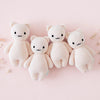 Four identical Cuddle + Kind Baby Kitten (Blush) dolls, hand-knit with black eyes and small smiles, are lined up side by side on a light pink background with a few small dried flowers scattered around.