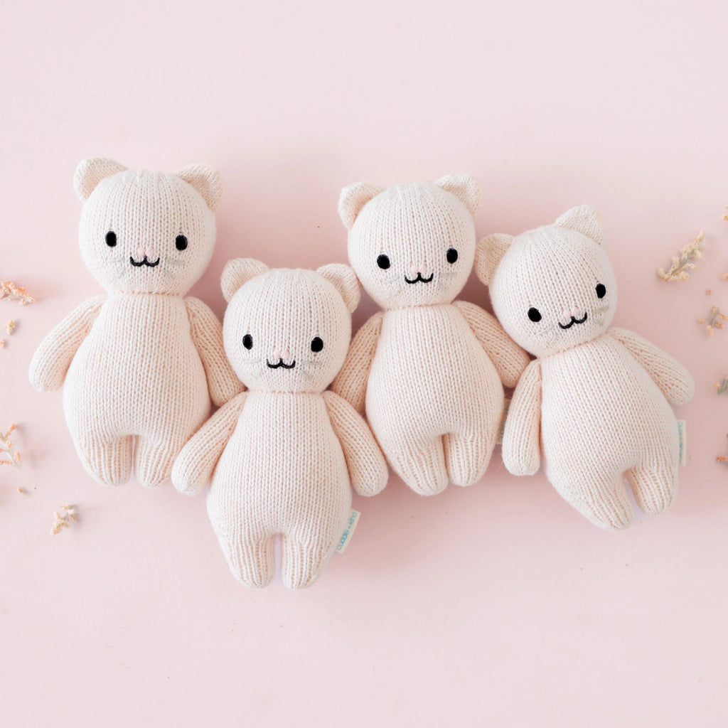 Four identical Cuddle + Kind Baby Kitten (Blush) dolls, hand-knit with black eyes and small smiles, are lined up side by side on a light pink background with a few small dried flowers scattered around.