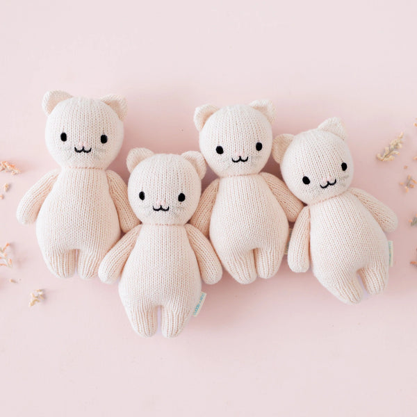 Four identical Cuddle + Kind Baby Kitten (Blush) dolls, hand-knit with black eyes and small smiles, are lined up side by side on a light pink background with a few small dried flowers scattered around.