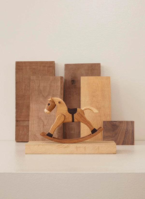 A small Spring Copenhagen The Rocking Horse displayed in front of various sized wooden planks arranged on a shelf. The background is softly lit, emphasizing the warm tones of the wood.