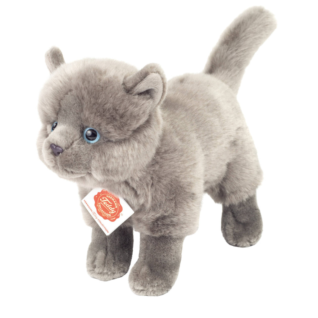 The Teddy Hermann Gray Standing Cat Stuffed Animal is a plush toy of a gray Carthusian cat with blue eyes, standing on four legs. This cat from the Hermann Teddy Collection features a soft, fluffy texture, a tag around its neck bearing the Steiff brand logo, and a slightly curled tail pointing upwards. Its child-friendly design makes it perfect for all ages.