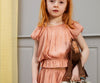 A young girl with long red hair stands indoors, clutching a brown stuffed horse in one hand and a small suitcase in the other. Dressed in a Maileg Princess Blouse - Melon (Final Sale) against a gray, paneled wall, she seems to have just emerged from a magical wardrobe, stepping straight into her fairytale dream.