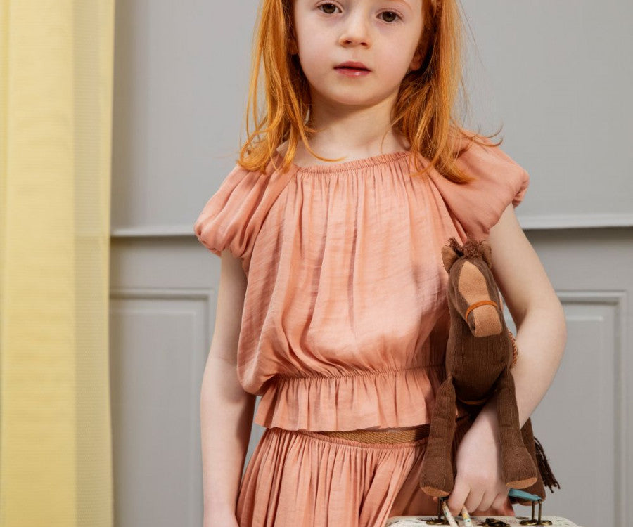 A young girl with long red hair stands indoors, clutching a brown stuffed horse in one hand and a small suitcase in the other. Dressed in a Maileg Princess Blouse - Melon (Final Sale) against a gray, paneled wall, she seems to have just emerged from a magical wardrobe, stepping straight into her fairytale dream.