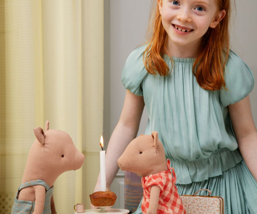 A young girl with red hair smiles while standing behind a table that has two stuffed pig toys, dressed in Maileg Princess Blouse - Mint (Final Sale), seated in chairs. In front of them is a small cake with a lit candle. The setting looks festive and cheerful, like a fairytale dream.