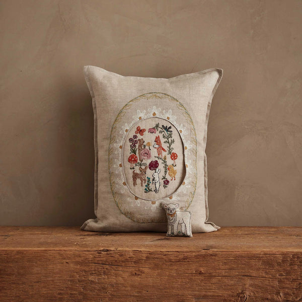 The Coral & Tusk Sugar Egg Pocket Pillow, featuring intricate, colorful embroidery of animals, flowers, and plants, rests on a wooden surface. In front is a small embroidered lamb plush—ideal for charming Easter decor.