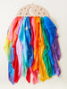 A colorful array of silk play scarves hanging from a Sarah's Silk Large Star Playsilk Display hanger shaped like a crescent moon with star cutouts. The scarves display a vibrant rainbow spectrum.