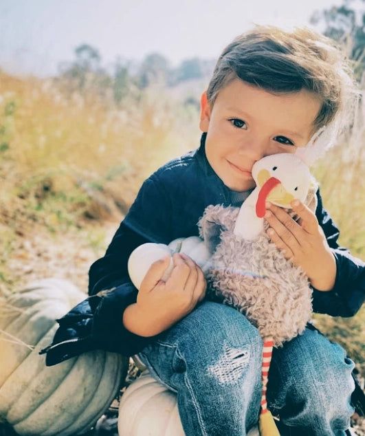 A young boy smiling gently, holding The Gracious Gobbler Bundle: Children's Book, Plush and Cards, sitting outdoors with a natural, blurred background that highlights his joy and the softness of the toy.