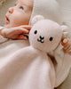 A baby in a light knit hooded outfit lies on a soft surface, holding the Cuddle + Kind Organic Cotton Baby Bear Lovey (Blush)—an ideal fair trade gift for any new arrival.