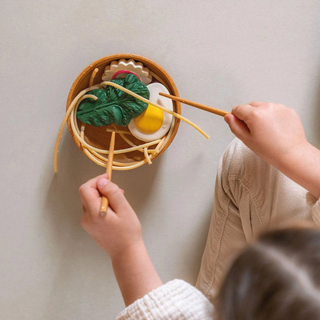 A child sits cross-legged on the floor, seen from above, while holding chopsticks and playing with a Ramen Bowl Playset (ships in approximately one week) made from natural non-toxic materials. The sensory play food includes plastic noodles, a green vegetable, a yellow egg, and other colorful toy food items in a brown bowl.