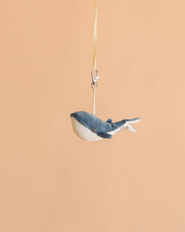 The Bukowski - Bubbles Whale Bag Charm features a small plush whale with a blue and white body, hanging from a beige ribbon and silver clasp against a plain peach background—making it the perfect cute accessory or mini bag charm.