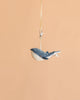 The Bukowski - Bubbles Whale Bag Charm features a small plush whale with a blue and white body, hanging from a beige ribbon and silver clasp against a plain peach background—making it the perfect cute accessory or mini bag charm.