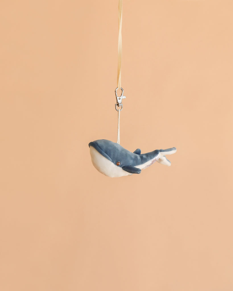 The Bukowski - Bubbles Whale Bag Charm features a small plush whale with a blue and white body, hanging from a beige ribbon and silver clasp against a plain peach background—making it the perfect cute accessory or mini bag charm.