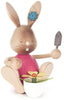 Collectible Dregeno Easter Figure - Rabbit Gardener With Flower Pot with articulated limbs, holding a small spade and a potted flower. Made in Germany, the bunny has a brown body, pink torso, and a blue flower decoration on