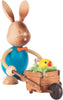 A Collectible Dregeno Easter Figure - Rabbit with Wheelbarrow from the Erzgebirge region, featuring a blue torso and an orange bow tie, is pulling a small cart containing a bright yellow chick and some greenery.