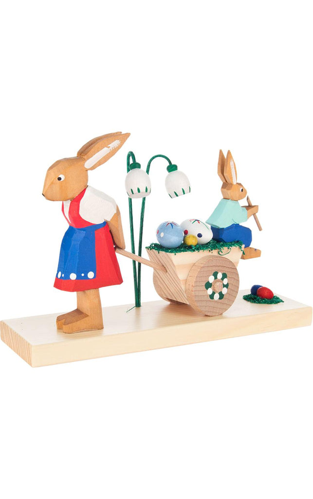 A handcrafted figurine from the Erzgebirge region of the Collectible Dregeno Easter Figures - Rabbit Mother and Son, featuring a mother rabbit pushing a cart full of colorful Easter eggs and wearing a red and blue dress, on a simple base against.