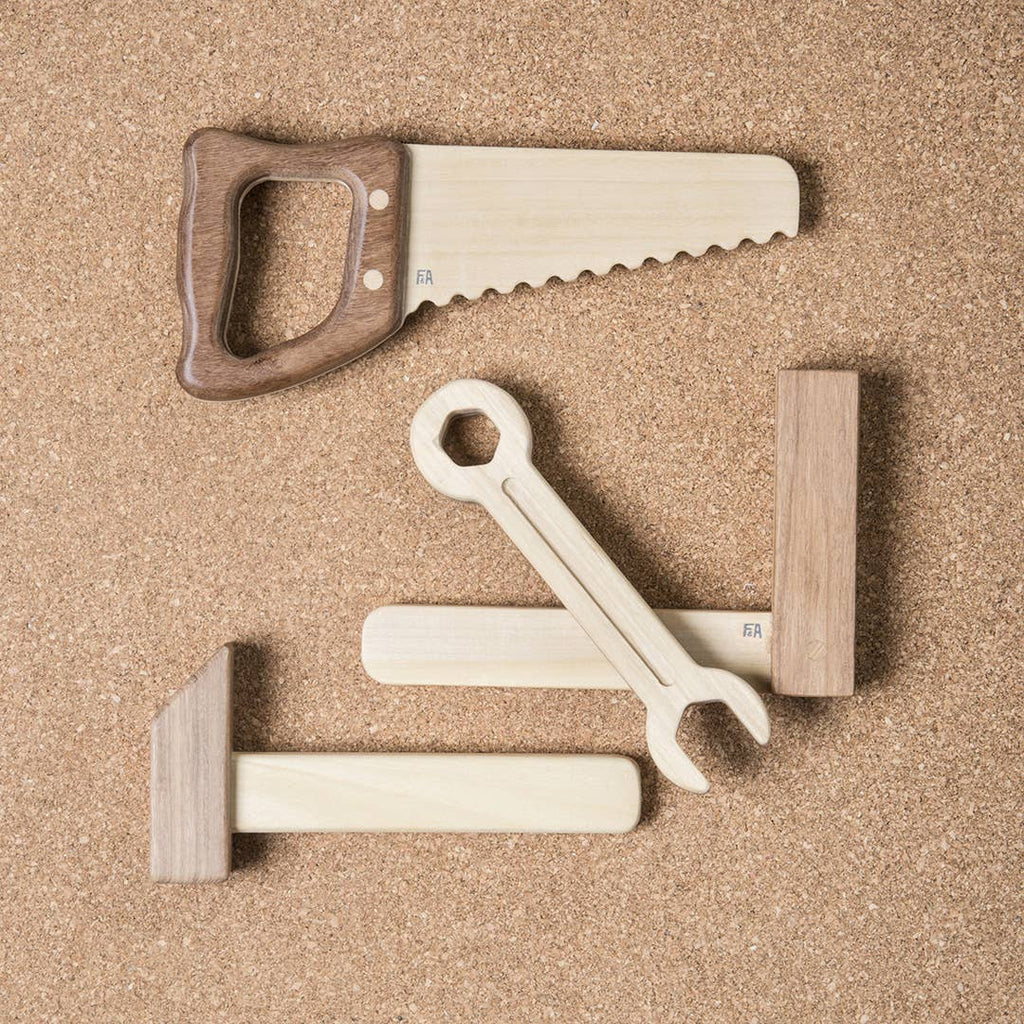 The Wooden Tool Set includes a saw, wrench, hammer, and mallet made from solid rubber wood and Sapele teak. Resting on a corkboard background, these toys showcase light and dark tones with clean, minimalist designs.