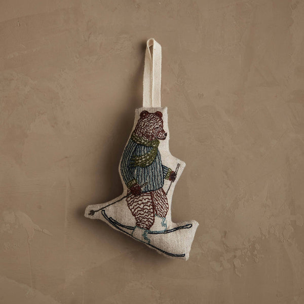 The Coral & Tusk Downhill Bear Ornament features a skiing bear in a striped sweater and scarf with poles, adding a playful winter touch as it hangs by a loop against a textured beige wall.