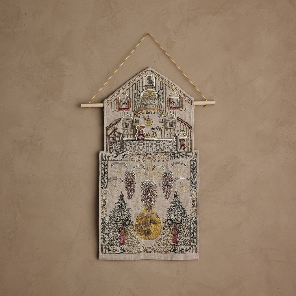The Coral & Tusk Christmas Clock Advent Calendar is a decorative textile wall hanging shaped like a cuckoo clock, with embroidered trees, pine cones, and red-clad figures on a beige textured background.