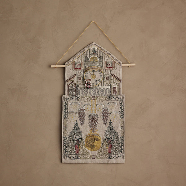The Coral & Tusk Christmas Clock Advent Calendar is a decorative textile wall hanging shaped like a cuckoo clock, with embroidered trees, pine cones, and red-clad figures on a beige textured background.
