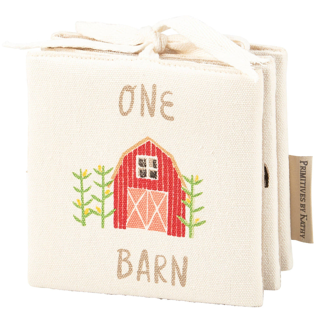 The Little Farm Soft Book is a soft infant book with a cream cover featuring a red barn and green plants, embroidered with the title "ONE BARN." Finished with a ribbon at the top, it’s a delightful farm-themed gift.