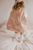 A child wearing the Cape - Pink sequins and white socks stands on a bed with light bedding. The photo, taken from behind, shows only the lower half of the child.