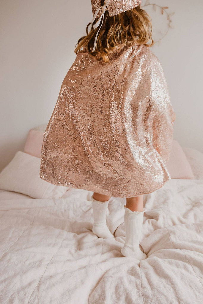 A child wearing the Cape - Pink sequins and white socks stands on a bed with light bedding. The photo, taken from behind, shows only the lower half of the child.