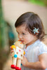 A young child with a flower in her hair wears a white shirt outdoors while holding and biting the Build-A-Robot toy, which features interchangeable heads for teaching emotions through play.