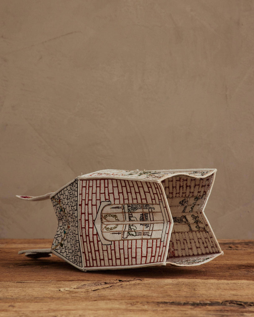 The Coral & Tusk Home for the Holidays Tissue Box Cover, shaped like a house with brick patterns and windows, rests on its side on a wooden surface against a plain beige wall.