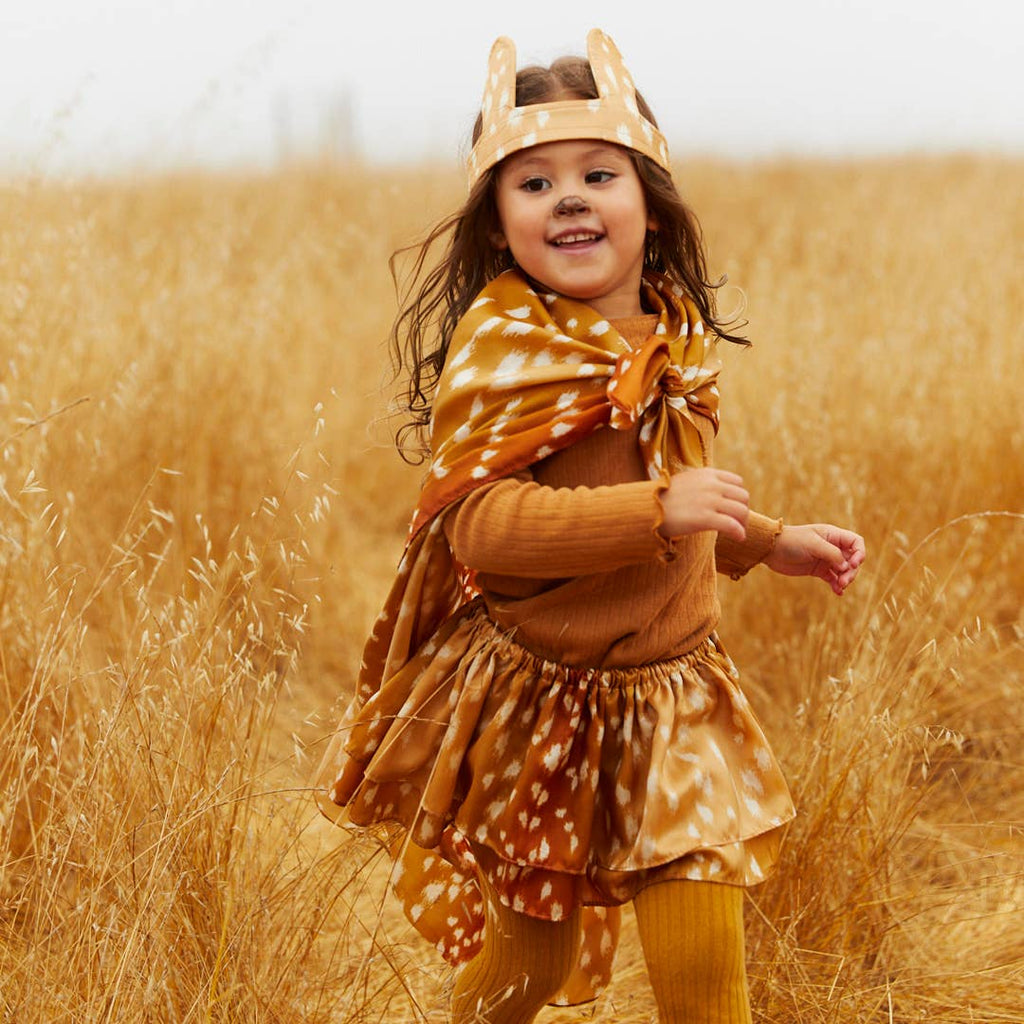 A young child wearing a Sarah's Silks Animal Print Playsilk - Fawn as a cape and skirt with an antler headband joyfully runs through a golden field of tall grass.