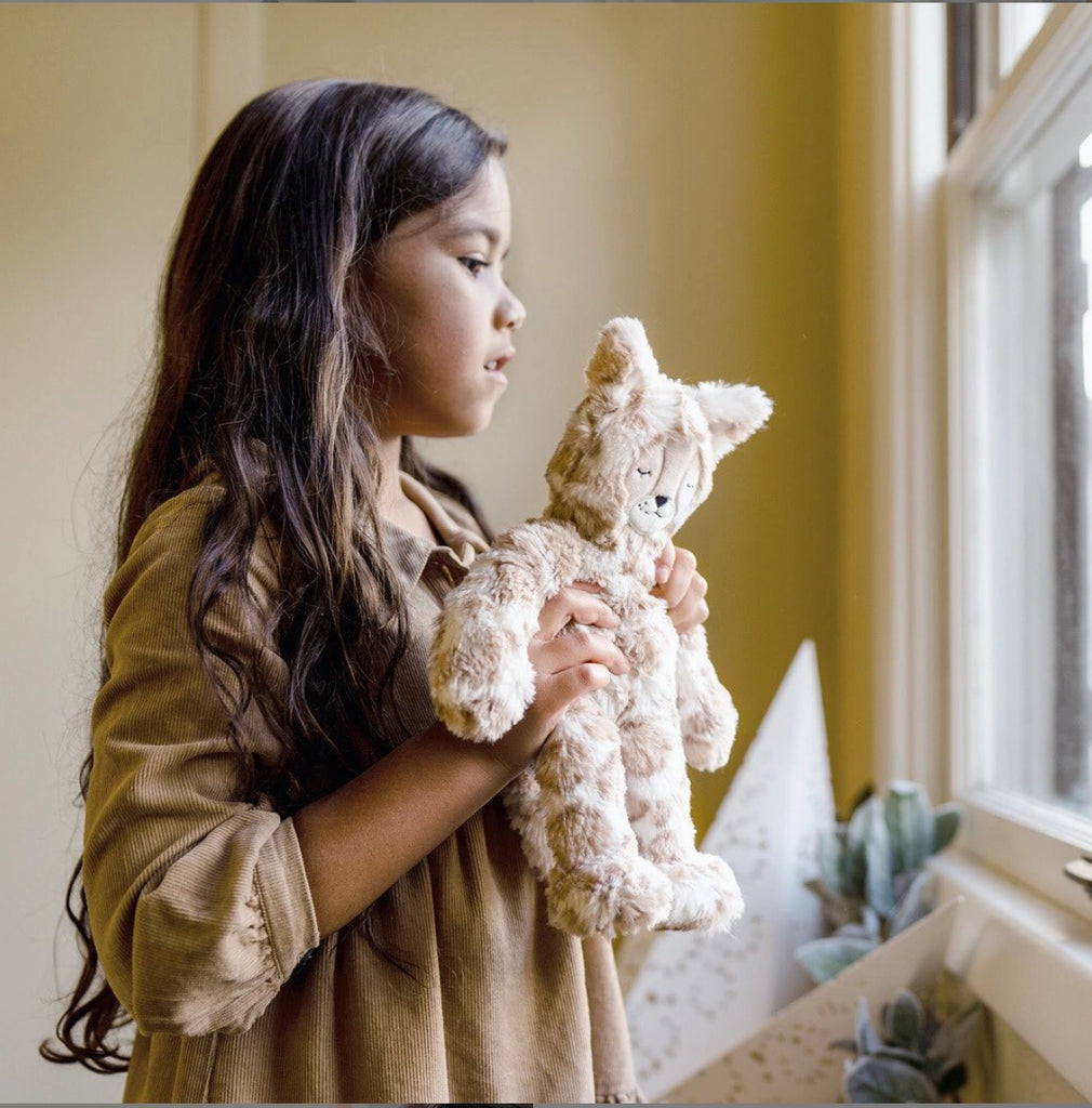 A young girl in a beige dress tenderly holds a Slumberkins Lynx Kin, gazing thoughtfully out a window in a room with yellow walls.