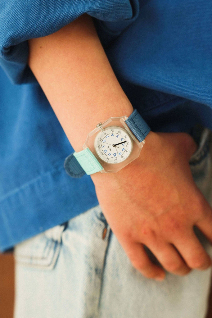 A person wearing a blue long-sleeve shirt and light denim jeans has a Mini Kyomo Watch - Blue Ocean with a translucent case, blue and light blue strap, black numbers, Japanese movement, and blue hands on their wrist.