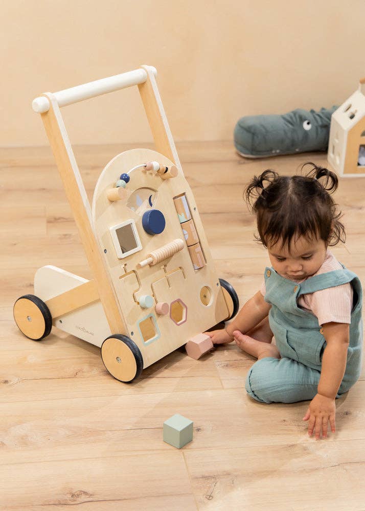 A toddler sits on a wooden floor next to the Wooden Activity Walker, which features a variety of colorful shapes and toys. She is wearing blue overalls and a pink shirt, and is playing with pastel-colored sensory toys and blocks spread out on the floor.