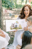 A woman in a white dress reads Winnie-the-Pooh: Finding Feelings to three young girls outdoors, surrounded by trees and festive bunting.
