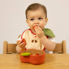 A baby wearing an apple bib sits at a wooden table, happily eating fruit from the Pepa the Apple Bowl & Spoon Set, a non-toxic baby tableware set with a red, apple-shaped bowl.