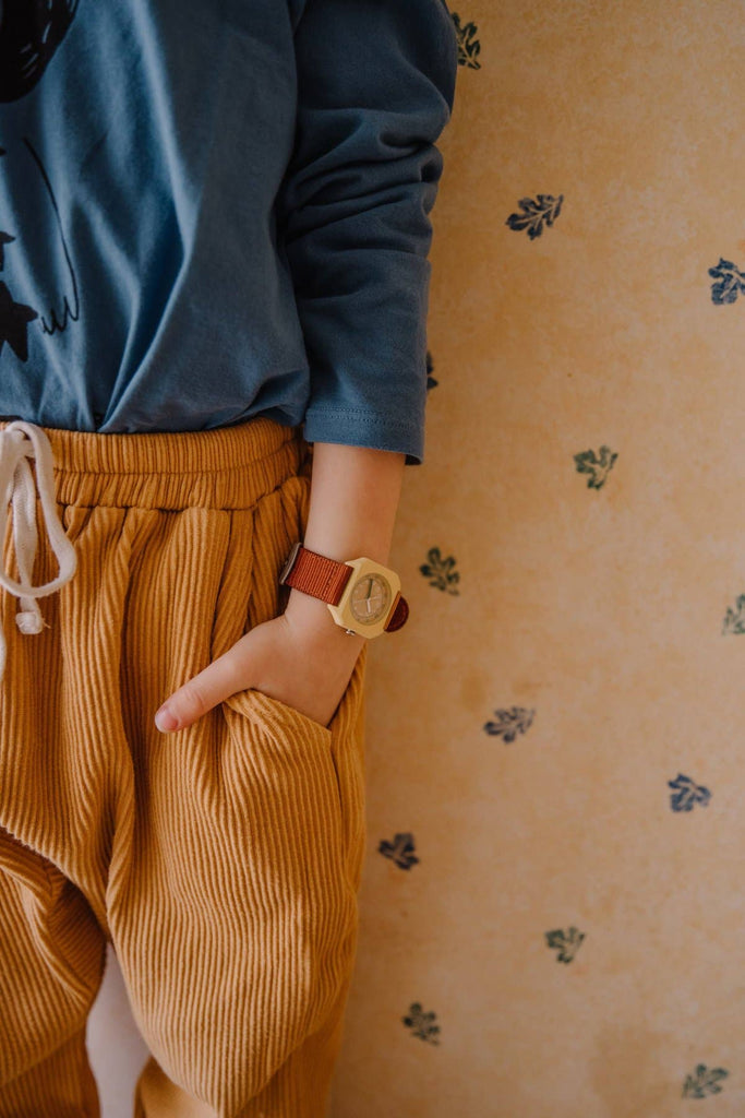 A person in a blue shirt and mustard corduroy pants stands with a hand in the pocket, showing off the Mini Kyomo Watch - Cherry Bomb with a brown strap, against floral-patterned wallpaper.