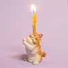 The Exotic Shorthair Cat Cake Topper features a small ceramic cat figurine in a red and white striped party hat, holding a yellow candle—perfect for topping cakes at cat-themed parties, shown against a light purple background.