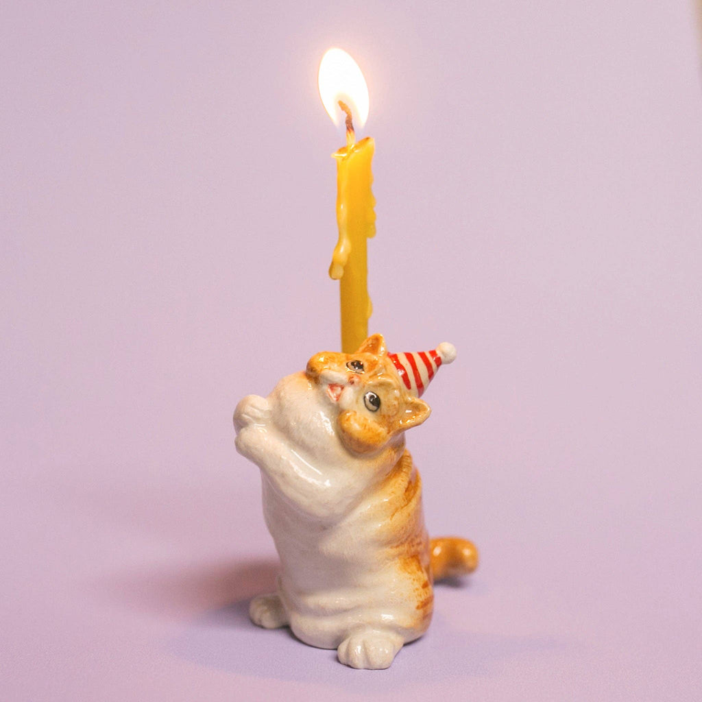 The Exotic Shorthair Cat Cake Topper features a small ceramic cat figurine in a red and white striped party hat, holding a yellow candle—perfect for topping cakes at cat-themed parties, shown against a light purple background.